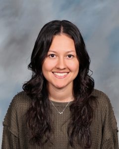 Smiling person with long dark hair wearing a brown sweater against blurry background